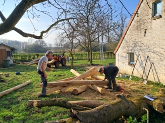 2 mannen bouwen mee aan de speelnatuur. Ze maken een klimbos van een aantal stammen die over elkaar gelegd worden en aan elkaar verbonden worden. Rechts de gevel van Vakantiehuis De Luysmolen, op de achtergrond enkele hoogstamfruitbomen met een ladder en een minitractor met frontlader. De tuin achter is begrensd met een haag en links zien we een stukje van het tuinhuis.