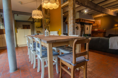 Zicht op de massieve eiken eetkamertafel met 8 stoelen errond. De stoel op de kop is bruin de andere zijn wit gelakt. We zien het keuken- en woongedeelte op de achtergrond. Boven de tafel branden 2 lampen.