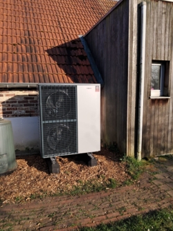 De warmtepomp tegen de gevel van het huis. Boven de warmtepomp zijn de dakpannen zichtbaar. Rechts ervan de uitsprong van het huis met houten gevelgekleding. Links een stuk van het compostvat.