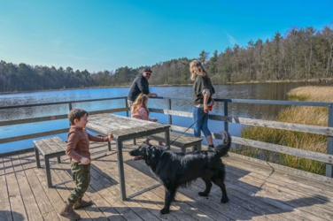 Een vader is met zijn twee kinderen op de steiger met picknickbanken en tafel aan de Ruiterskuilen op Donderslaghei. De jongen heeft veel aandacht voor onze Mazzel die samen met Helena ook op de steiger is.
