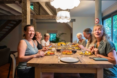 Een familie zit samen rond de eettafel. Het eten is geserveerd. Links in het donker het woongedeelte, rechts het raam met zicht op de tuin.
