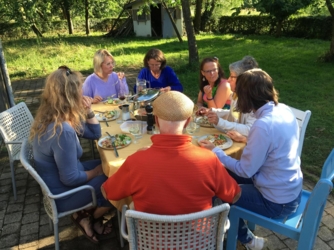 Een groep van 7 mensen zit rond de tuintafel in de tuin van vakantiehuis De Luysmolen in het Kempenbroek. Het eten is geserveerd. Op de achtergrond zien we in de tuin een aantal bomen en het tuinhuis.