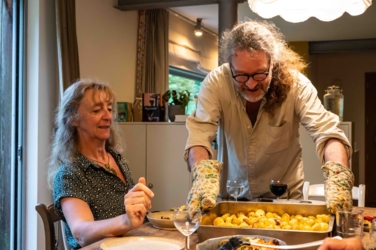 De kok van dienst zet een schotel op tafel. Helena links helt een beetje naar achter om de man plaats te geven. Boven tafel brand een lamp en op de achtergrond zien we het licht en het raam van de keuken.