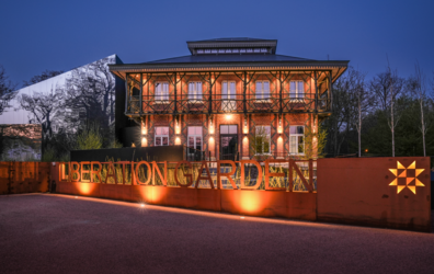 Een foto van het Museum Liberation Garden in Leopoldsburg genomen in de avond. Het gebouw is langs buiten helemaal verlicht. Aan de straat is er een afsluiting gemaakt van beton en daarboven de letters LIBERATION GARDEN helemaal doorzichtig. Ook deze afsluiting staat belicht met spots.